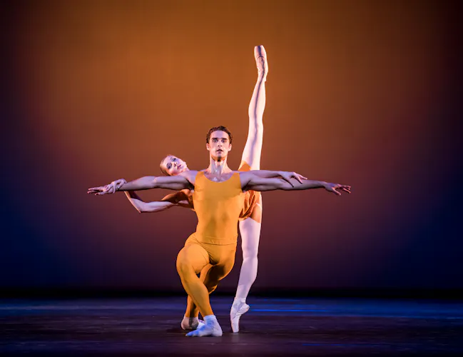 Two dancers wearing yellow leotards appear on stage. The dancer in front crouches with one knee bent in front of him and arms outstretched to the side. Behind him the other dancer has her legs in the splits in a vertical line from the floor, she balances her body on the man's back. (1 of 3)