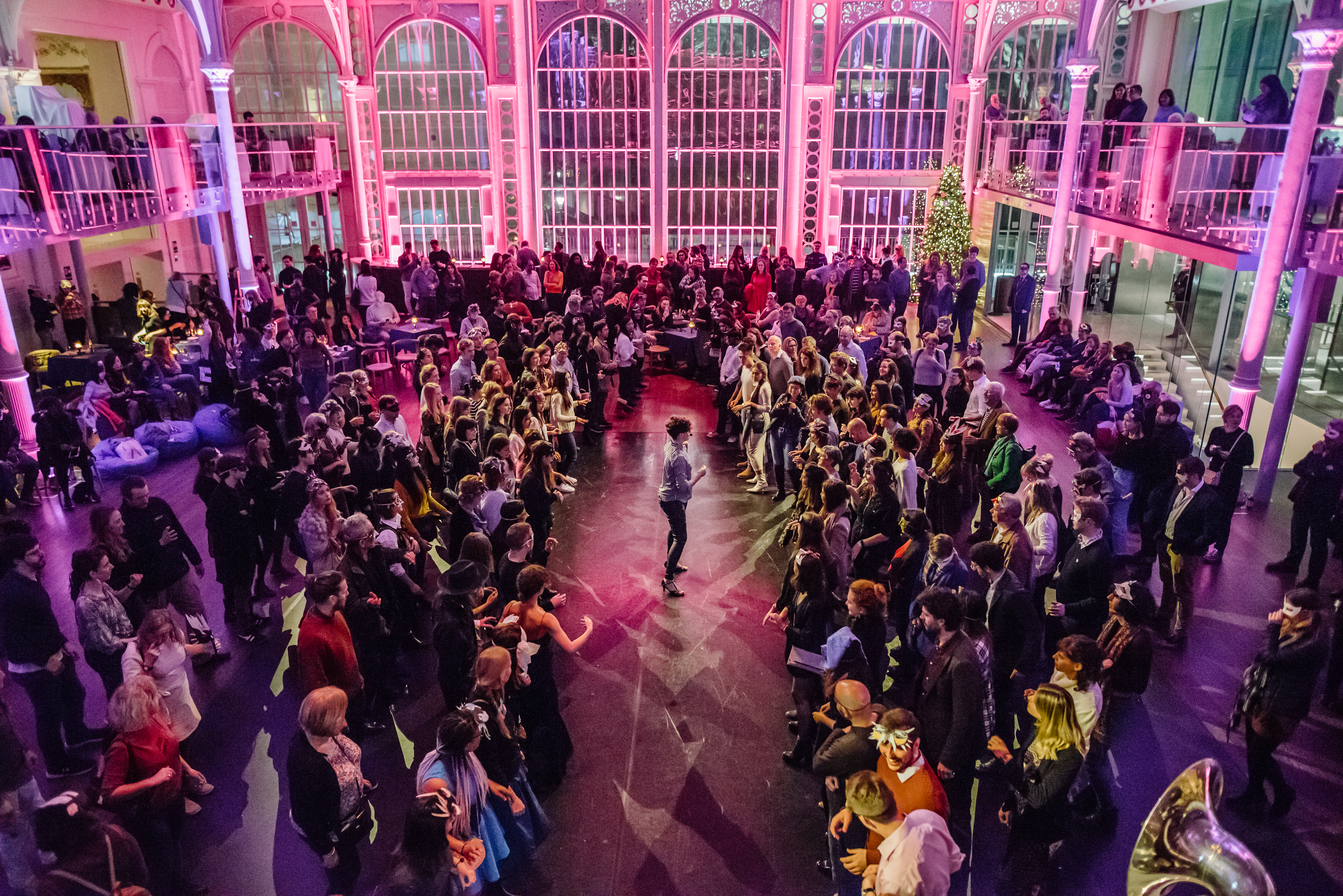 The Paul Hamlyn hall is filled with people. It is illuminated by purple lighting. In the centre a performer addresses the standing crowd.