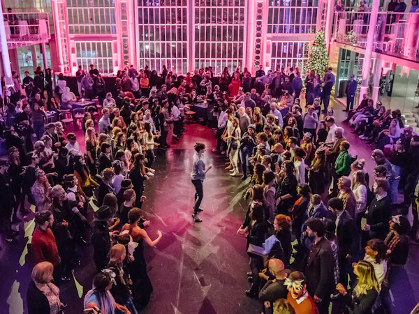 The Paul Hamlyn hall is filled with people. It is illuminated by purple lighting. In the centre a performer addresses the standing crowd.