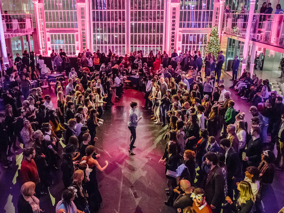 The Paul Hamlyn hall is filled with people. It is illuminated by purple lighting. In the centre a performer addresses the standing crowd.