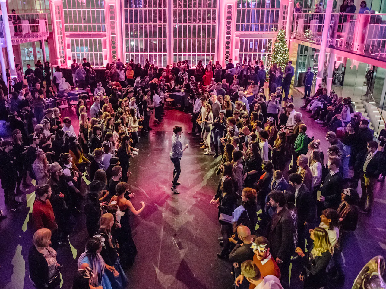 The Paul Hamlyn hall is filled with people. It is illuminated by purple lighting. In the centre a performer addresses the standing crowd.