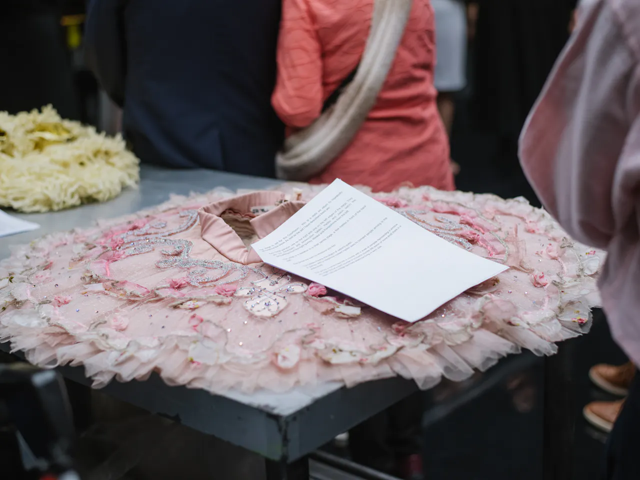 A sparkly pink tutu lies flat on a black table. A woman in a pink shirt stands behind the table.