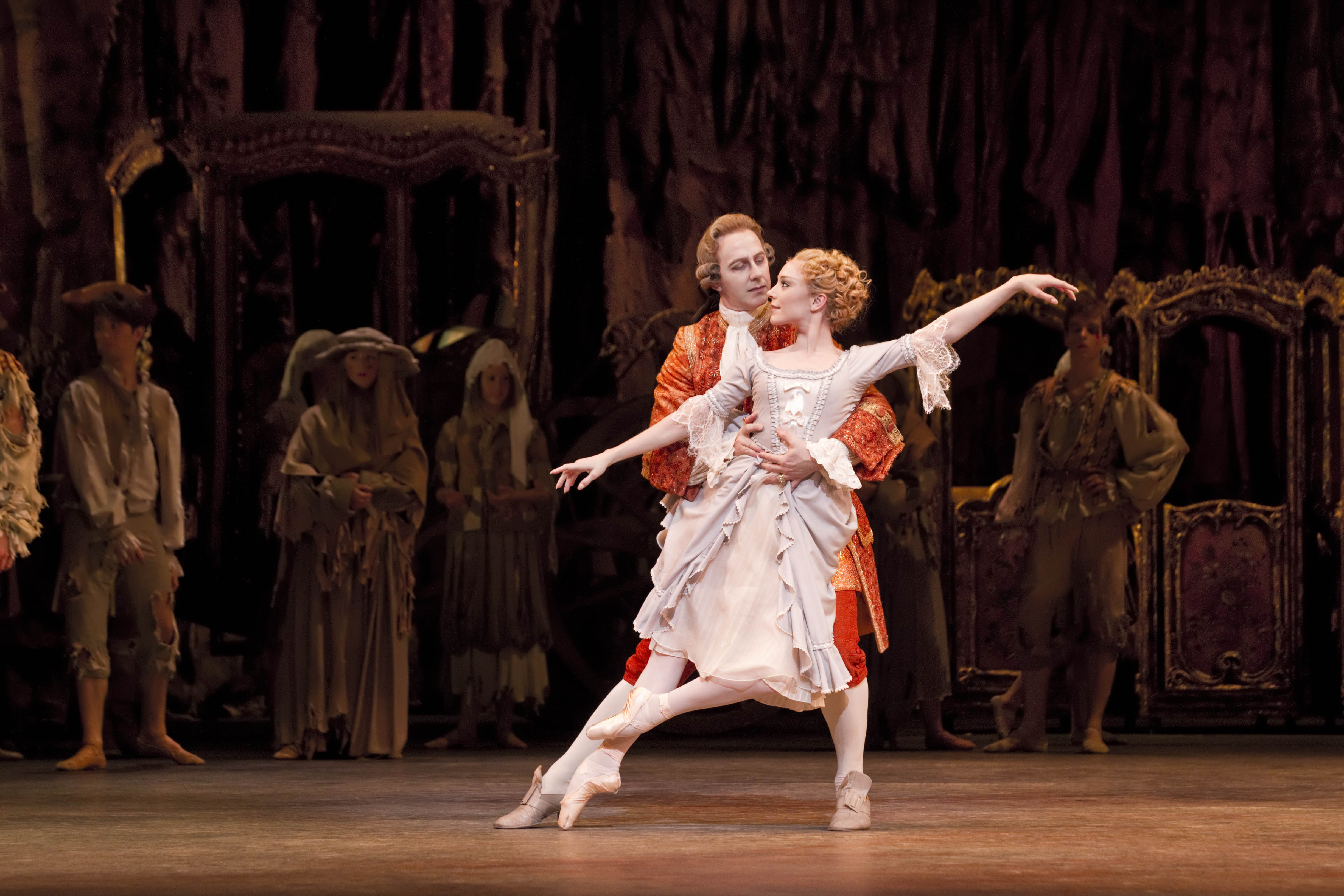 Two dancers appear on stage in period dress. The man holds the woman by the waist from behind, he is wearing a long red velvet jacket. The woman has both arms outstretched, and wears a pink and cream dress. (5 of 8)