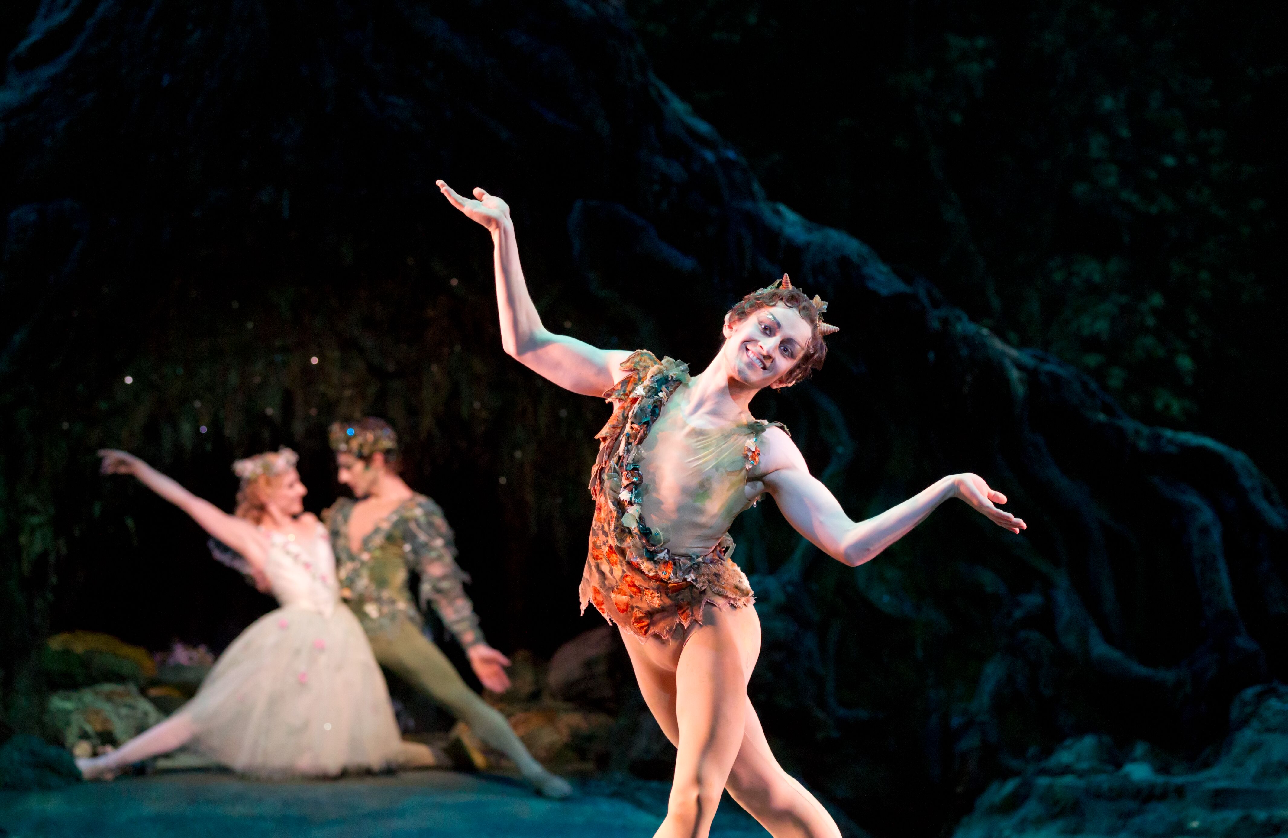 A man dressed as a fairy stands centre stage with his arms in a shrugging motion. Behind him are a male and female fairy kneeling closely together. (1 of 4)
