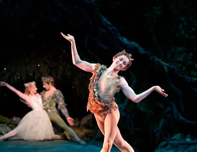 A man dressed as a fairy stands centre stage with his arms in a shrugging motion. Behind him are a male and female fairy kneeling closely together. (1 of 4)