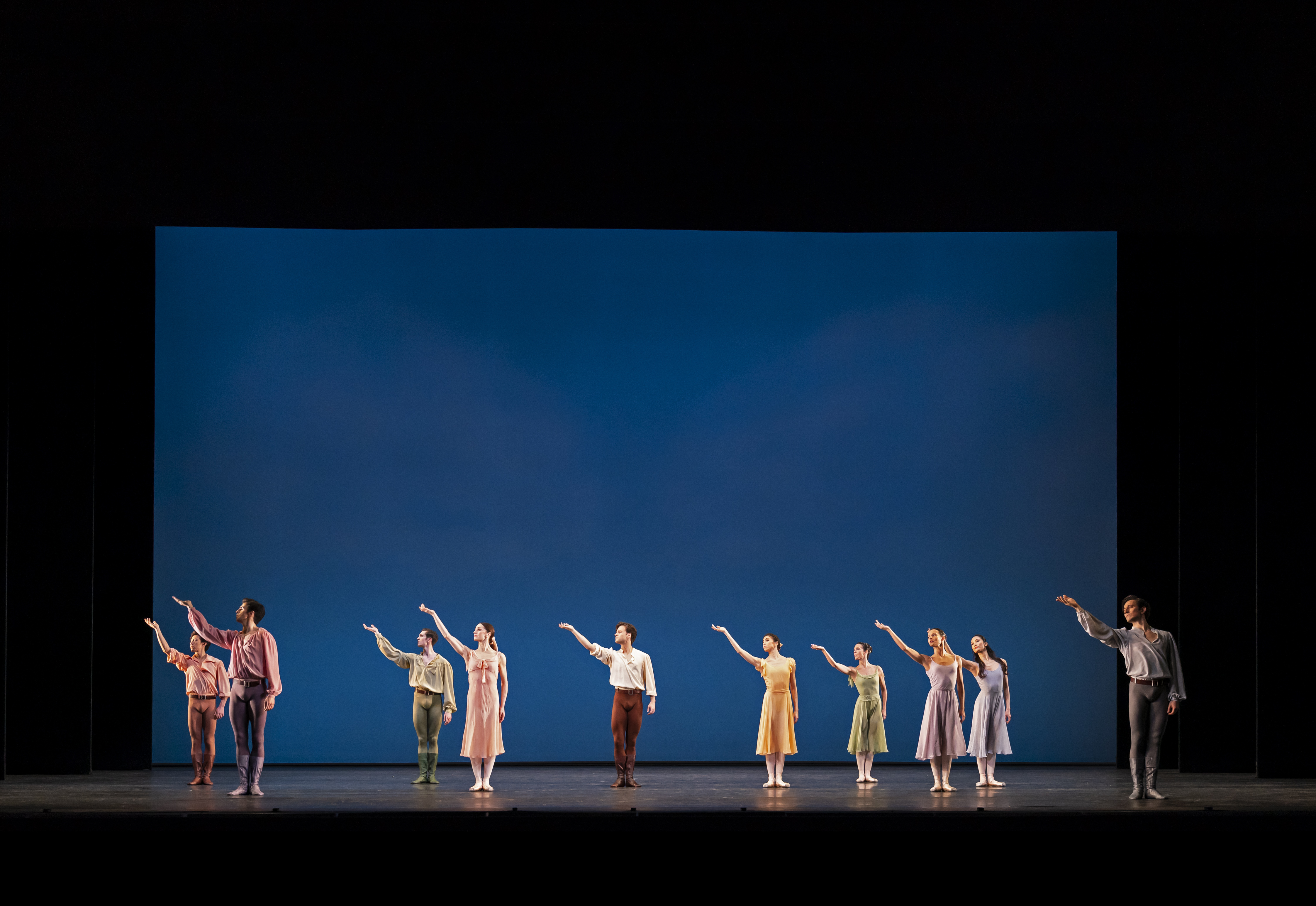 Ten dancers perform on an empty stage with a blue background. They are all facing the audience and all have their right arms raised. (8 of 8)