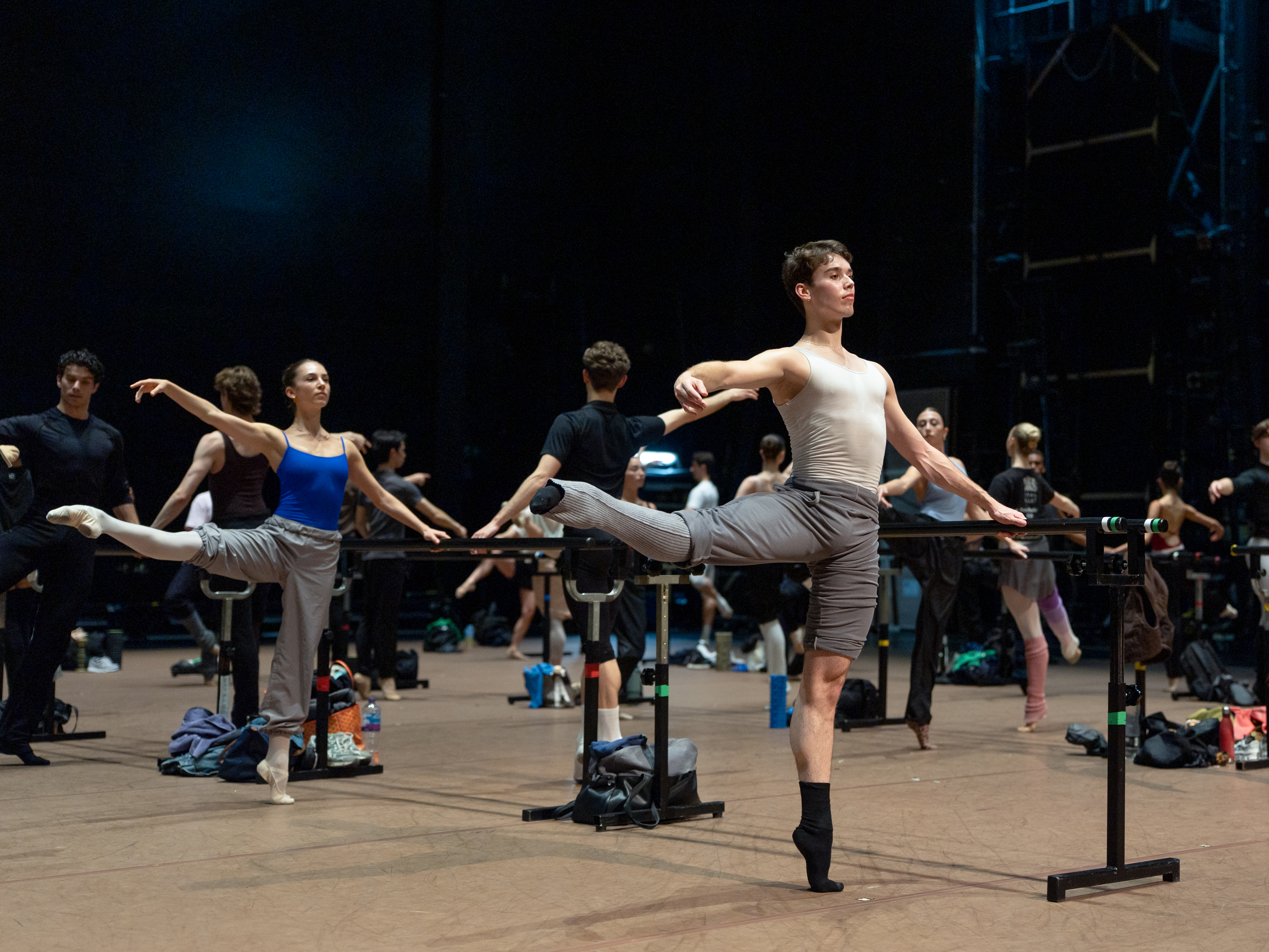 Ballet dancers stand on stage during class. They wear loose fitting trousers and stand on one leg, with the other leg raised to the side. They hold onto a ballet barre with their left hands. 