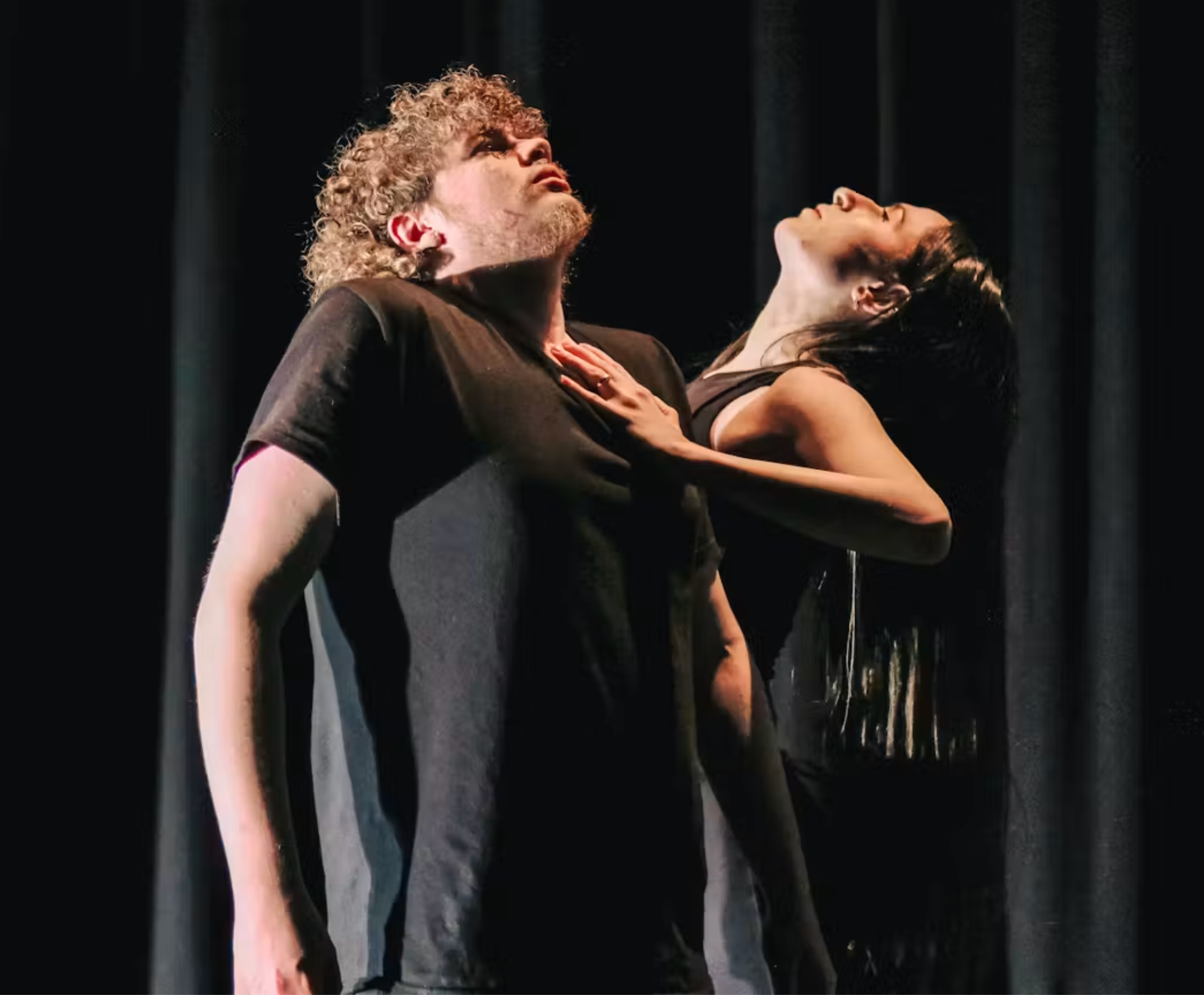 Two dancers stand in a dark room. One wear a black short sleeve tshirt and holds their shoulders up towards their neck looking upwards, the other places one hand on the others chest and looks up. 