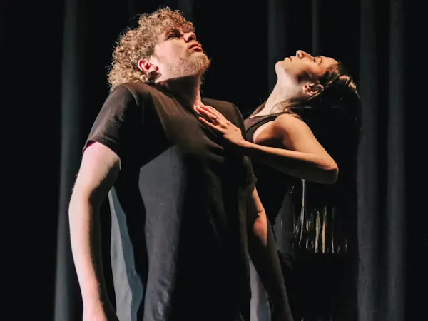 Two dancers stand in a dark room. One wear a black short sleeve tshirt and holds their shoulders up towards their neck looking upwards, the other places one hand on the others chest and looks up.