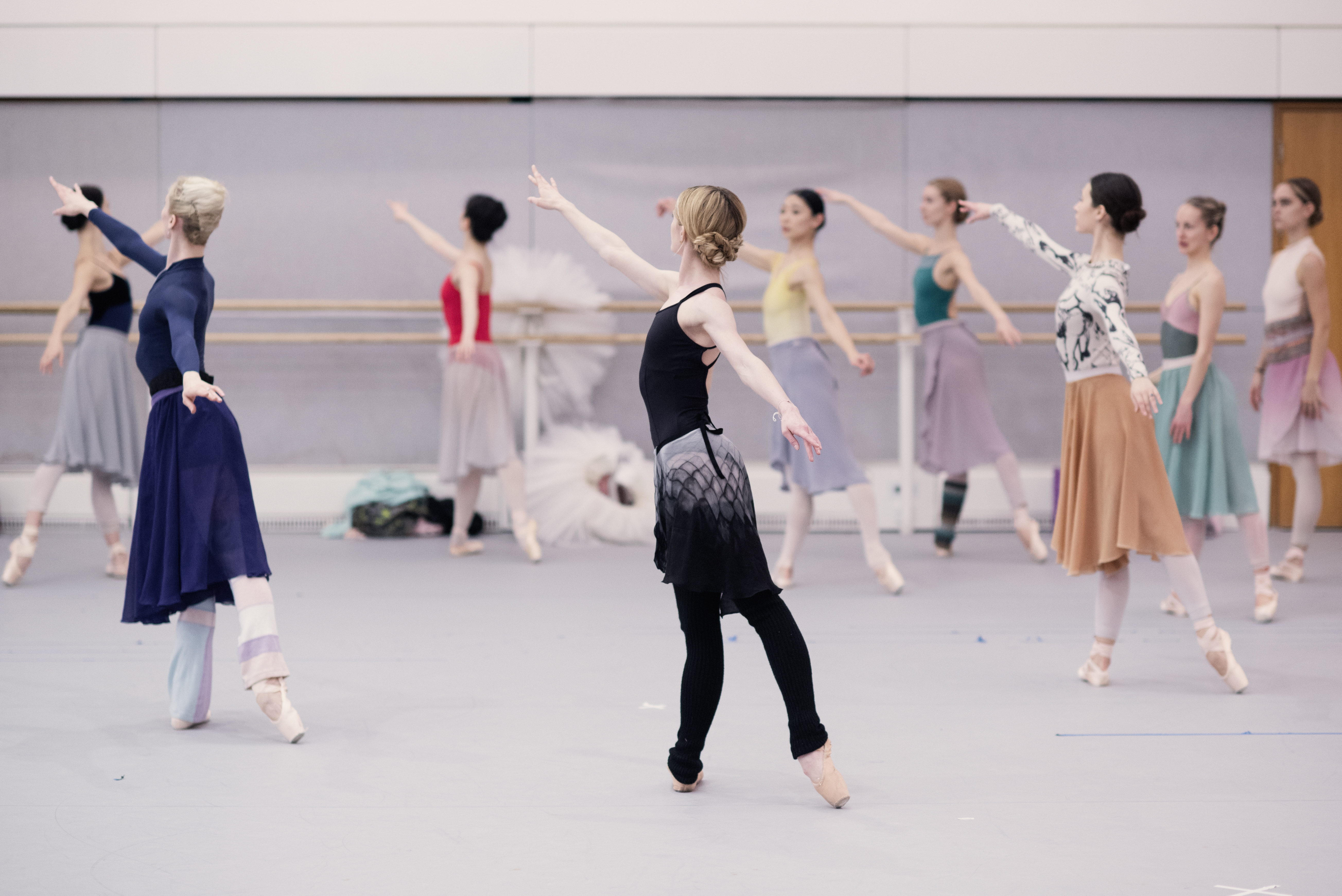 Dancers rehearse in a white studio. They stand with their backs to to the camera with arms raised.