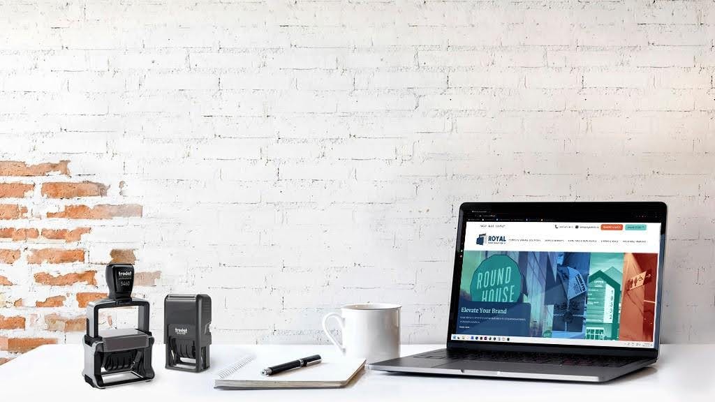 Desk in front of a white-painted brick wall. On the desk are two self-inking stamps, a notepad and pen, a coffee cup, and a laptop with the Royal Stamp website on the screen.