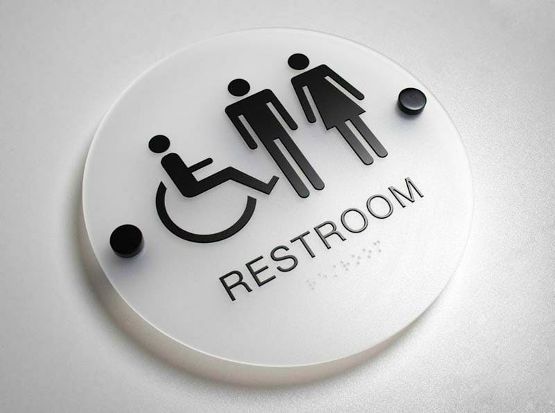 Circular sign with black graphic on frosted glass. Text reads 'RESTROOM'. Above the text is a graphic with three symbols for: wheelchair user, male, female.