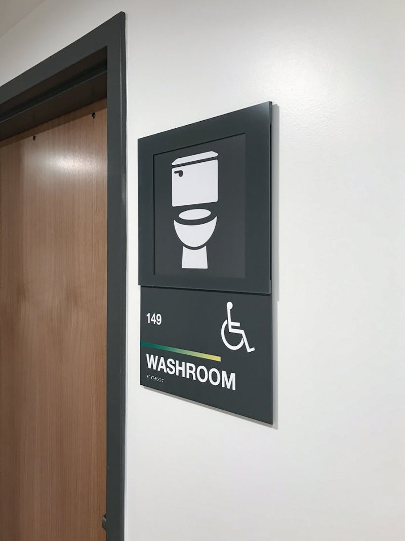 Side view of a door with a sign mounted on the right side. The sign is dark gray with white graphics. The graphic contains a toilet at the top; below that is the universal symbol for wheelchair user above the word 'WASHROOM'.