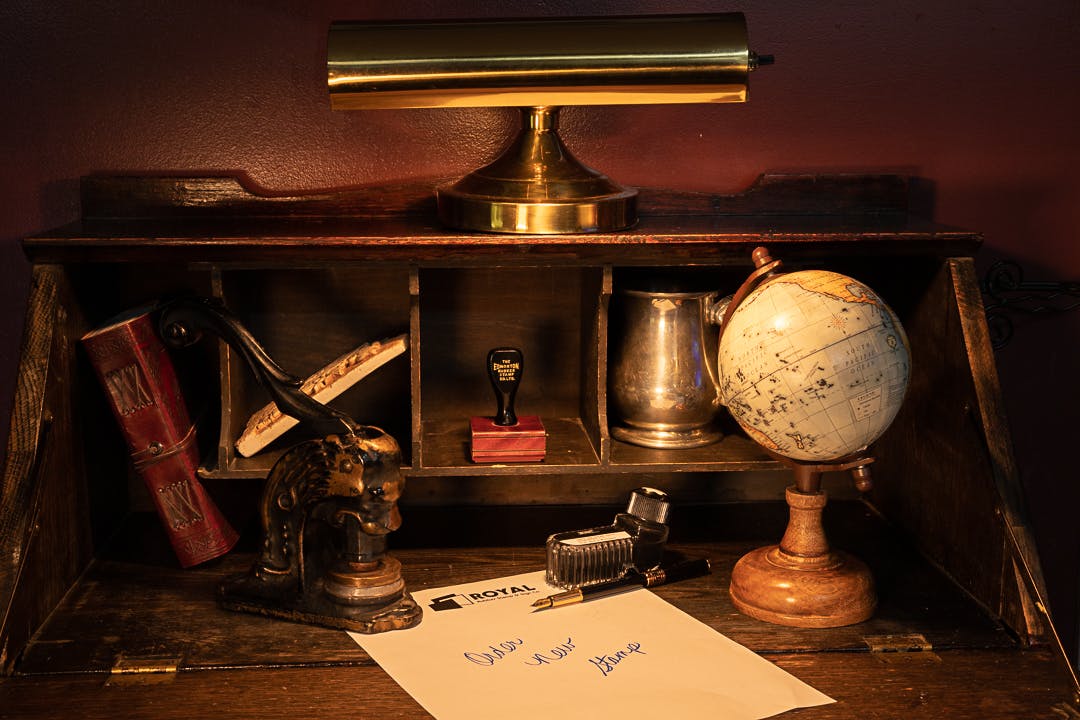 A vintage scene featuring a dark wooden desk illuminated from above. On the desk, there is a globe, books, and various accessories arranged. A piece of paper with the Royal Stamp logo is present, along with an old-style stamp and a modern stamp. Handwritten words on the paper read 'Order New Stamp'.