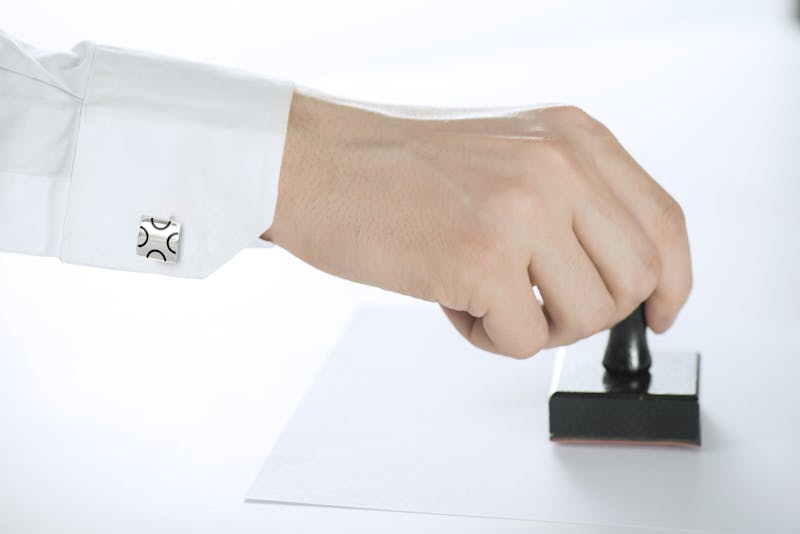 (Stock iamge) A hand, from the wrist down, stamping a piece of paper. The wrist has a cuff with silver cufflink.