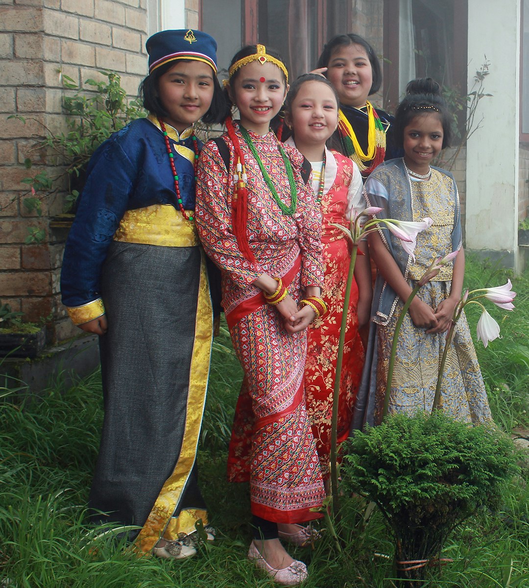 Celebrating Diversity: Five young students, representing different cultures and traditions, proudly showcase their vibrant traditional attire as they stand amidst the lush greenery and blooming flowers of our school.