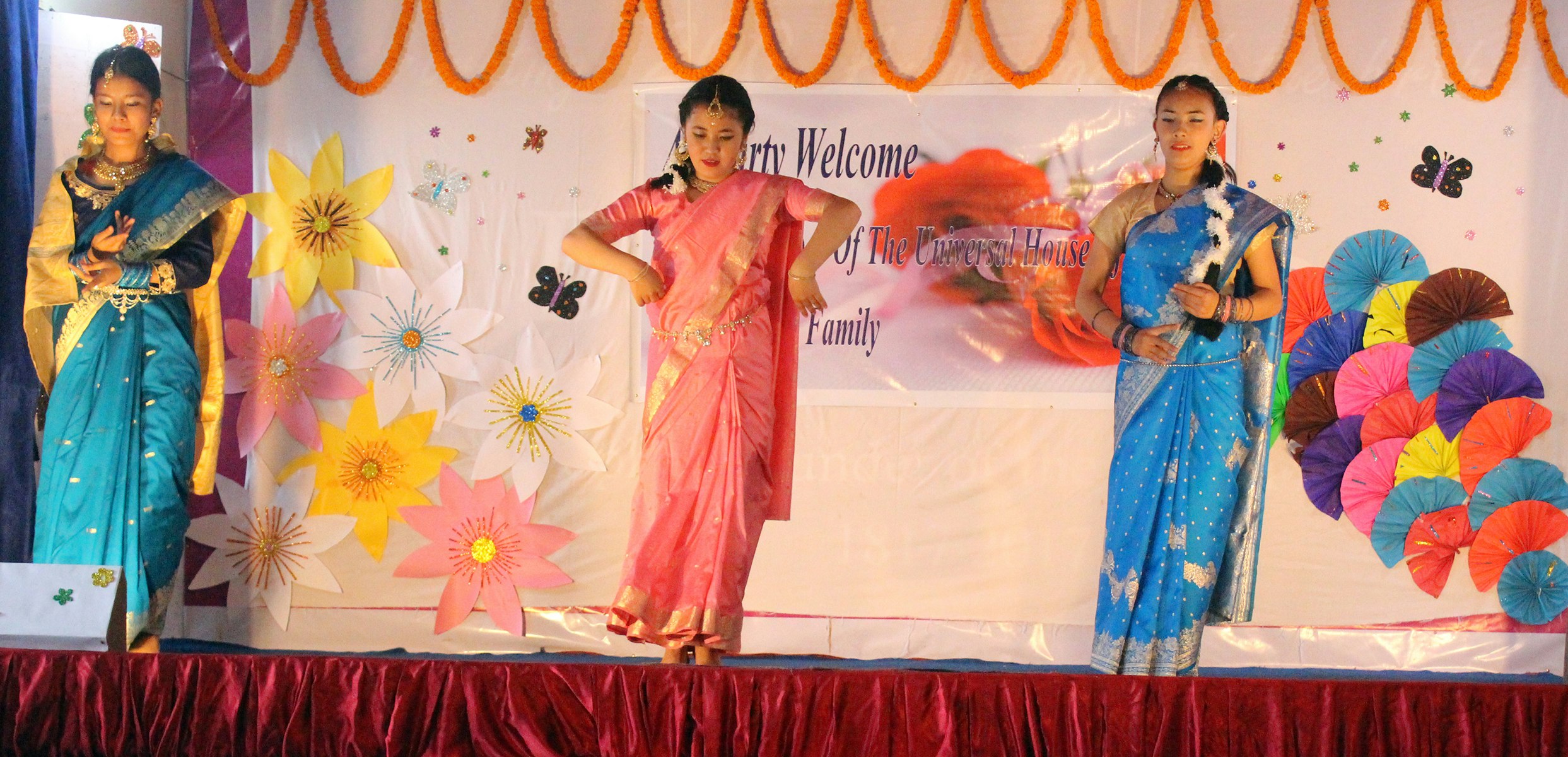 Graceful Rhythms: The stage comes alive as three talented girl students, adorned in vibrant blue and pink saris, captivate the audience with their mesmerizing dance performance on this special occasion.