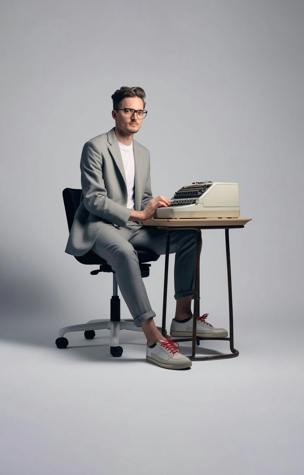 Man met bril in pak met sneakers zit aan een kleine tafel met oude typewriter.