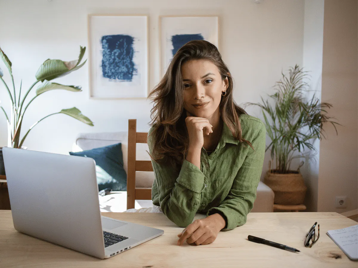 Een persoon zit aan een houten bureau met een laptop en een notitieboekje, met kamerplanten op de achtergrond.