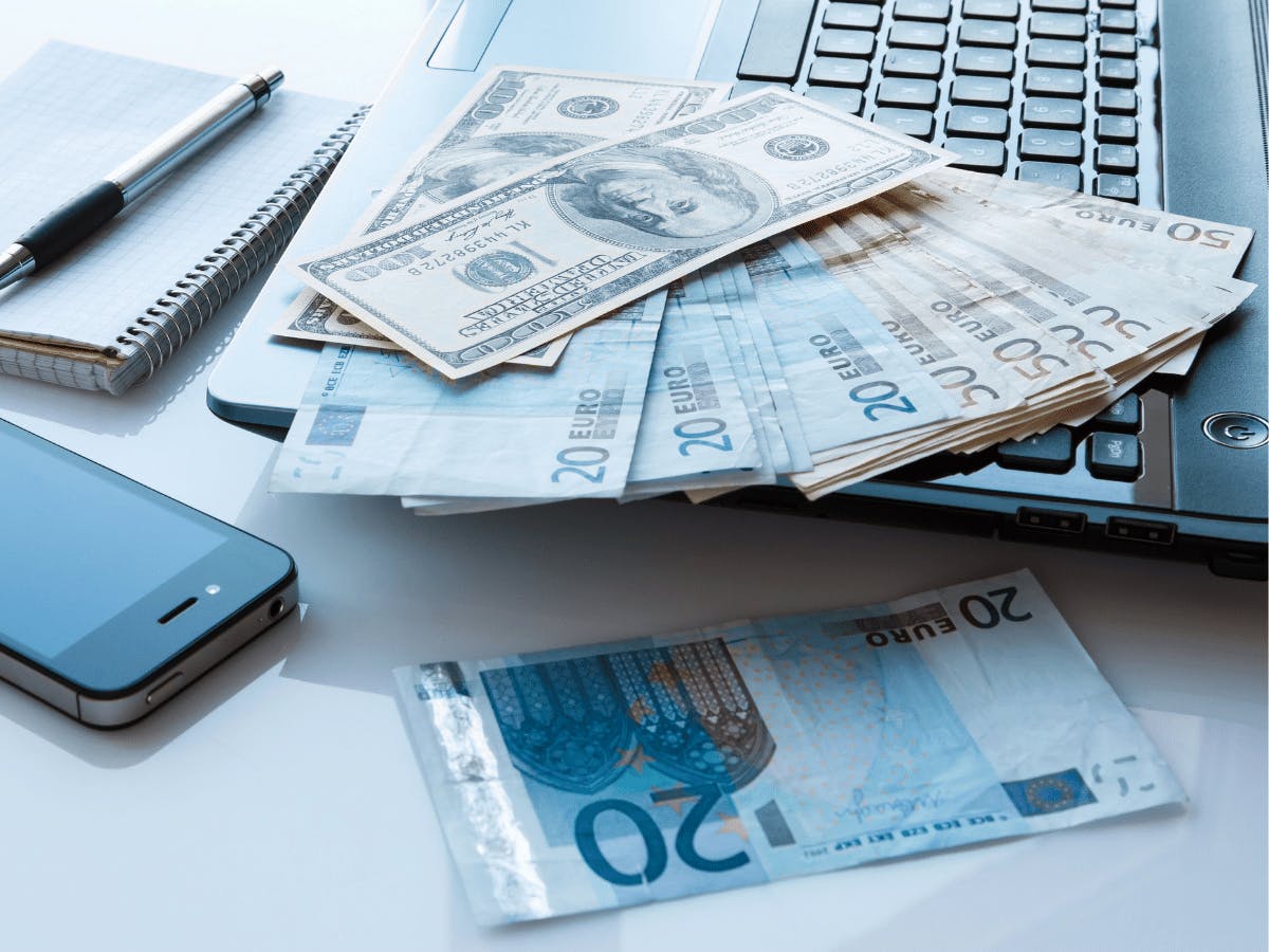 Een stapel euro en dollar biljetten liggen op een het toetsenbord van een laptop met ernaast een iPhone, kladblok en pen