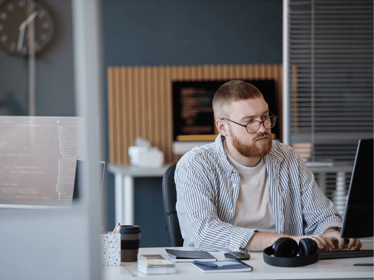 Een man met een bril en een baard zit achter een bureau met een koptelefoon op; hij typt op een computer terwijl hij naar een beeldscherm kijkt.