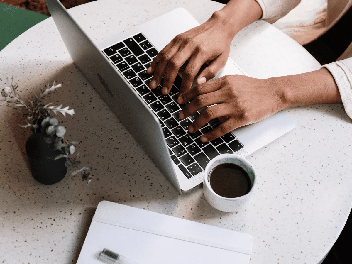 Iemand die aan een tafel zit te werken, typend op een open laptop met de handen boven het toetsenbord, met een mok koffie en een klein potplantje ernaast.