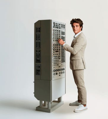 Een persoon in een pak staat naast een machine met heel veel knopjes.