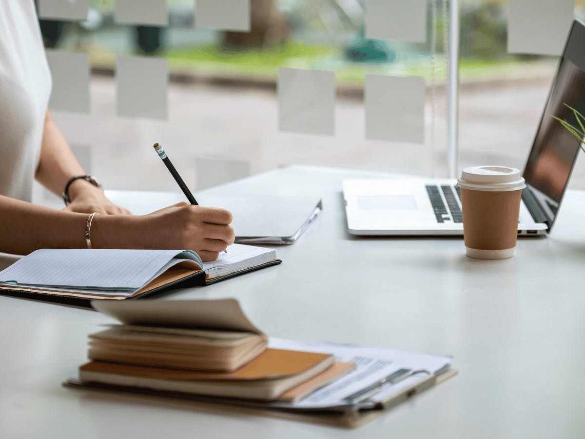 Vrouw schrijft in notitieboek met laptop en diverse kantoorartikelen op het bureau