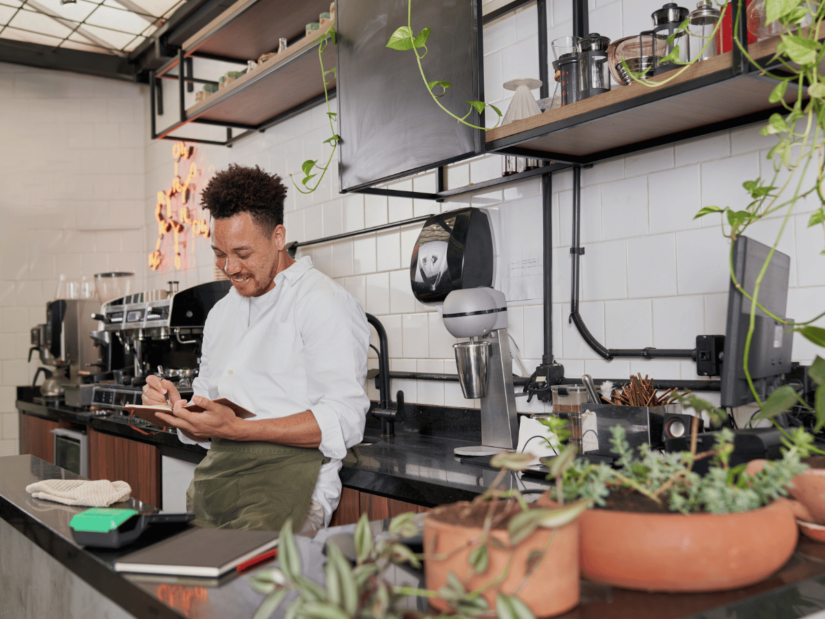 Een persoon die achter de balie in een koffiezaak staat en in een notitieboekje kijkt.
