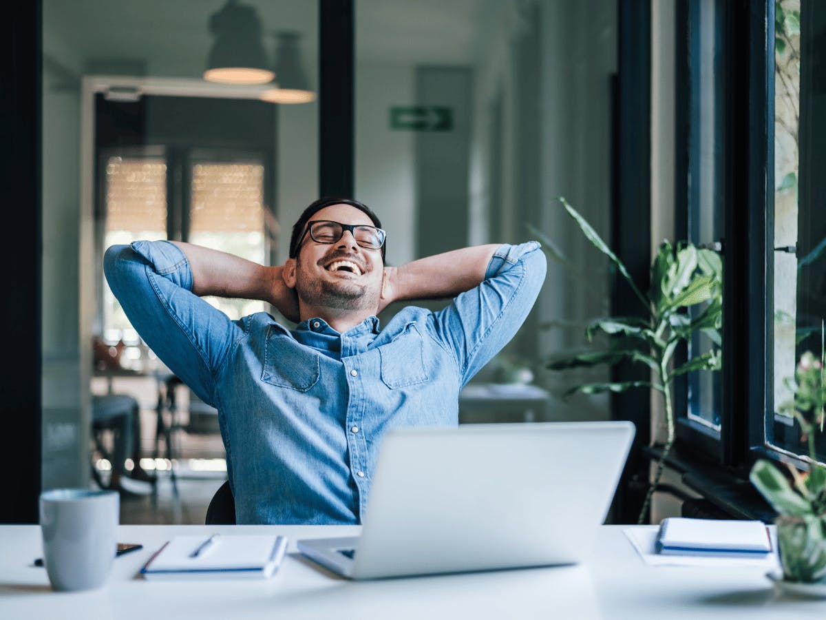 Een glimlachende persoon die aan zijn bureau zit met zijn armen achter zijn hoofd.