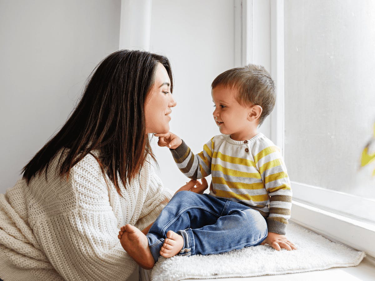 Een persoon in een witte trui zit naast een raam met een jong kind in een gestreept shirt en een spijkerbroek.