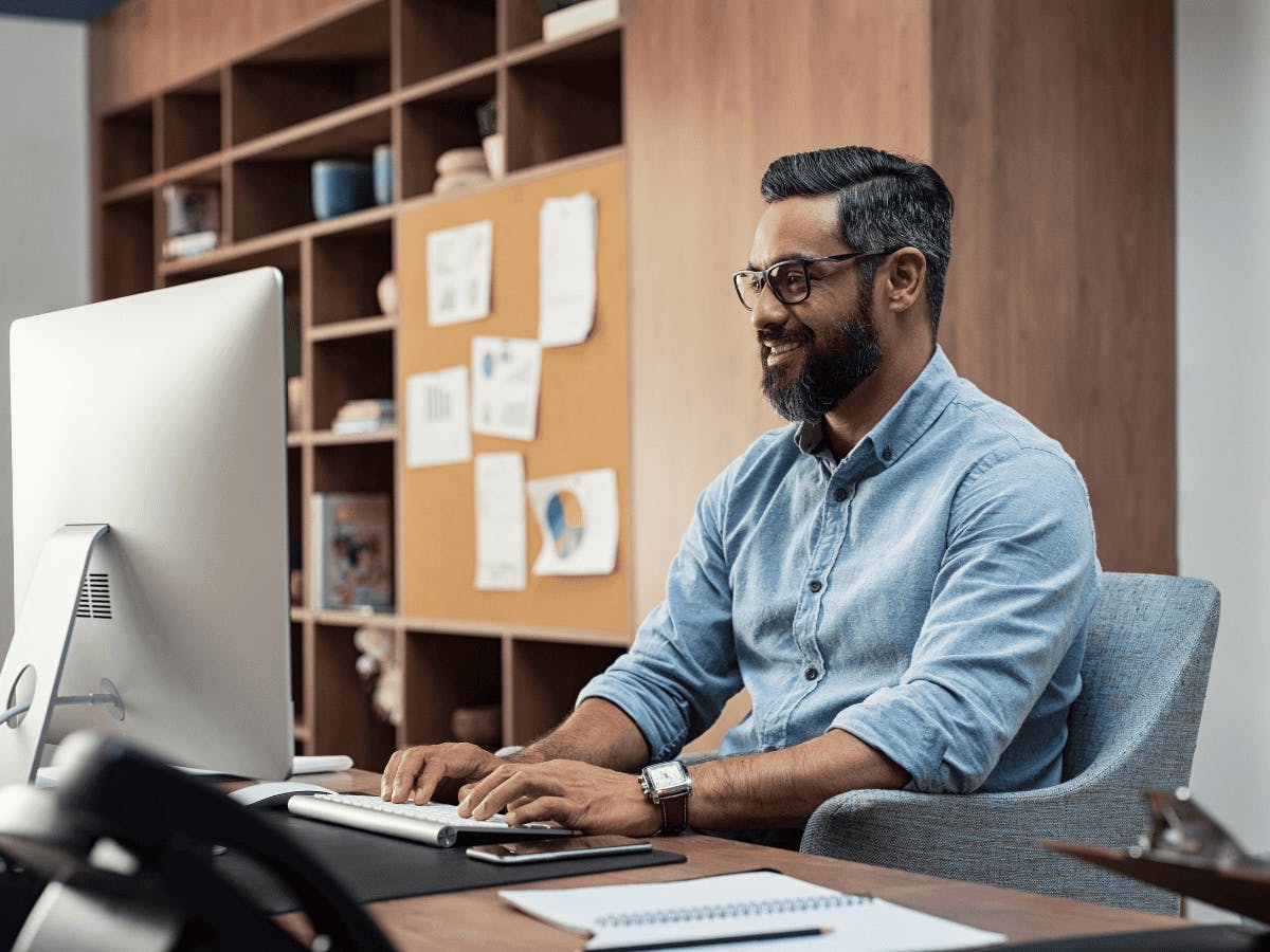 Een man met een bril en een blauw shirt zit aan een bureau en typt op een toetsenbord voor een computer.