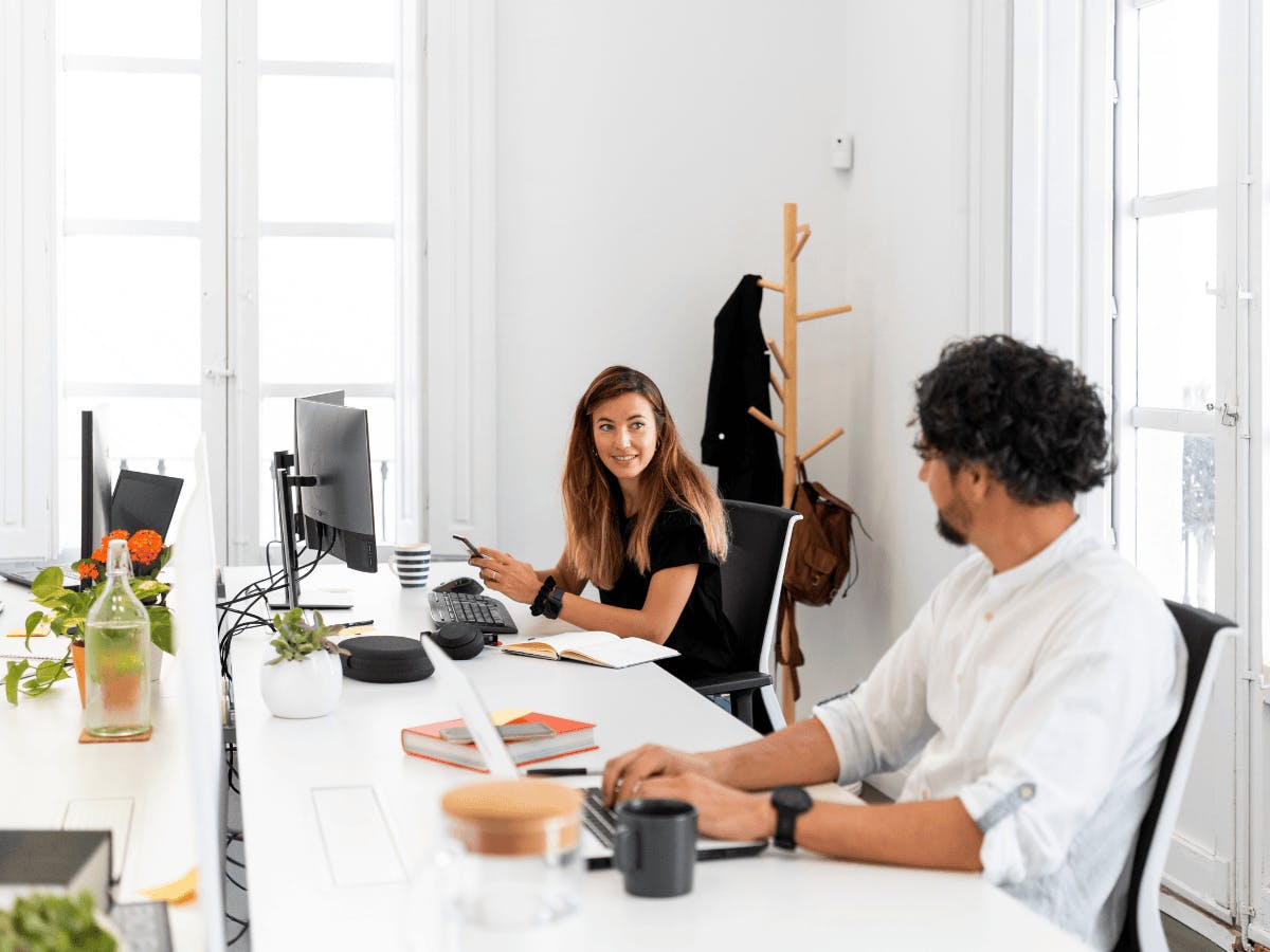 Twee mensen zitten in een licht kantoor aan een bureau en praten met elkaar.