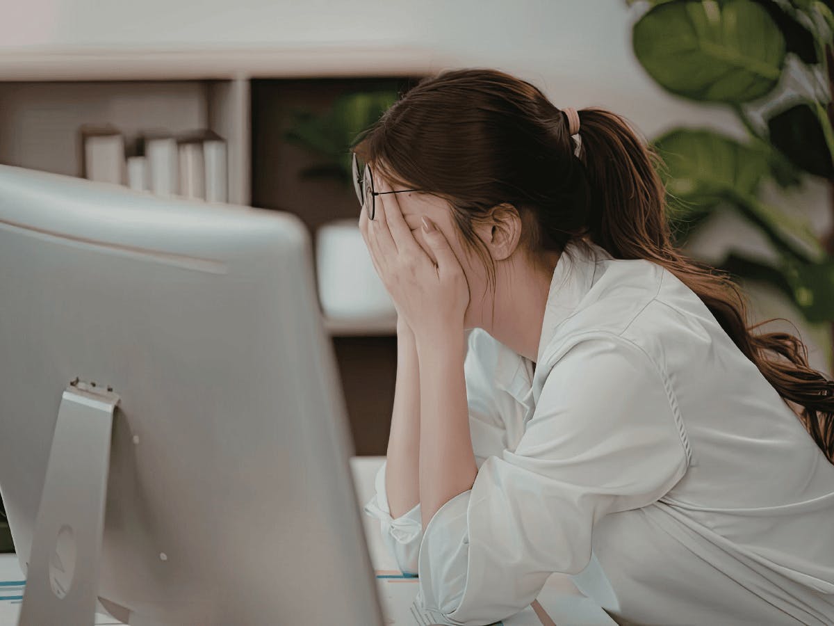 Een persoon met een bril die zijn gezicht met zijn handen bedekt, zittend voor een computerscherm in een kantooromgeving.