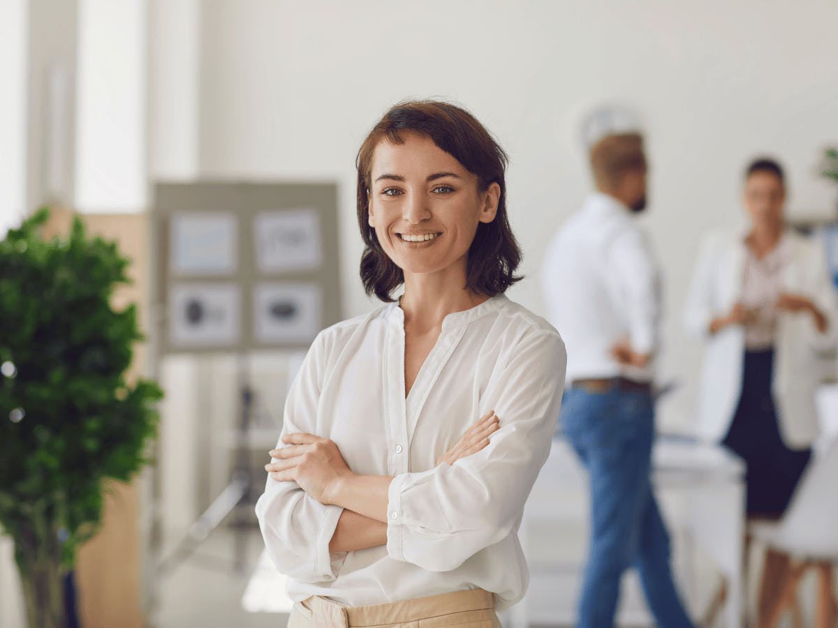 Een glimlachende persoon met gekruiste armen staat voor een wazige kantoorfacade met collega's.