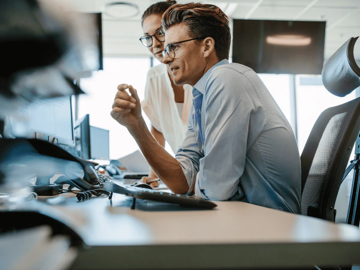 Twee collega’s die samen aan een bureau werken en naar hun computerscherm kijken.
