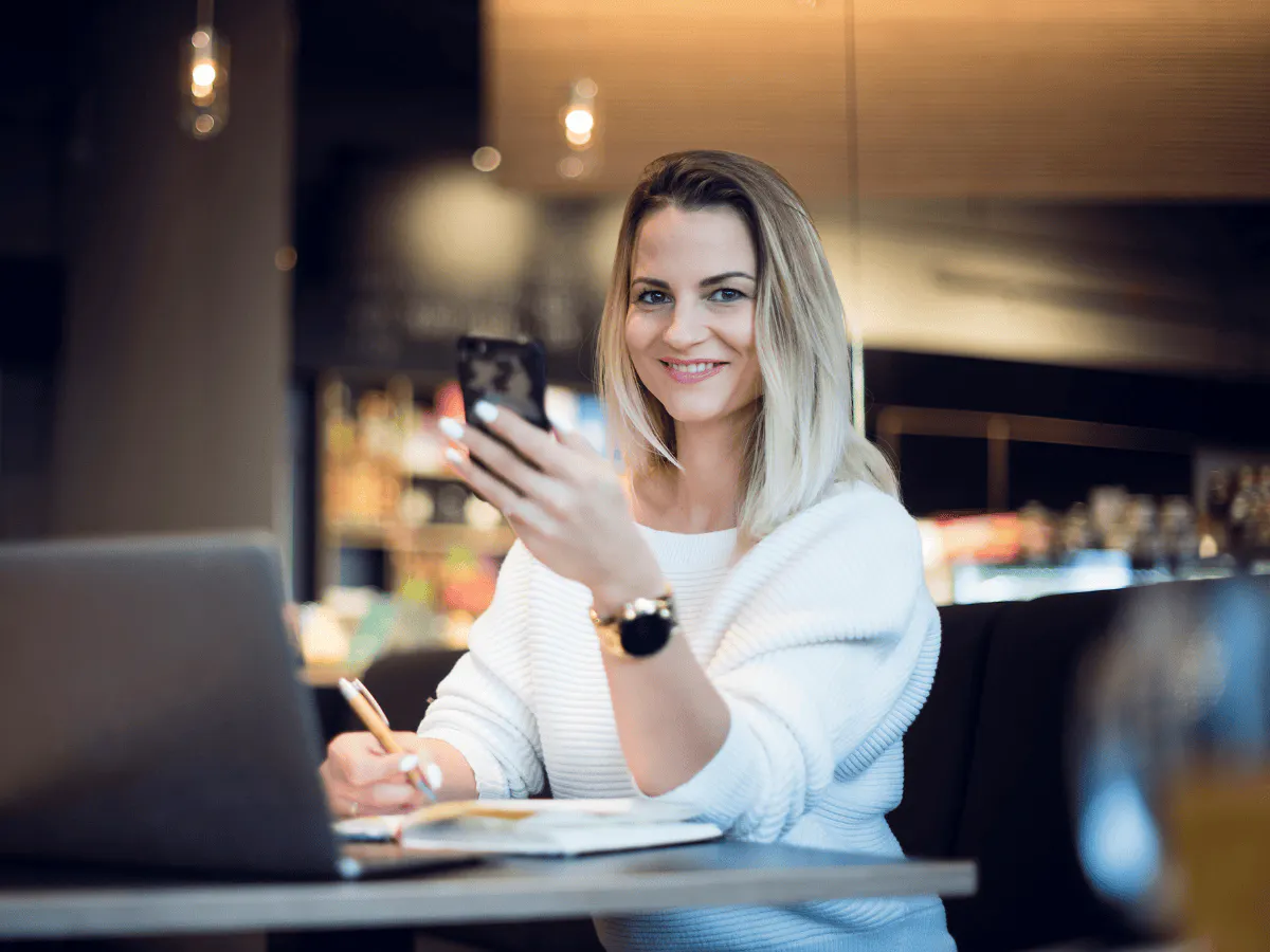 Er zit iemand aan een tafel met een laptop en een telefoon, terwijl zij in een notitieboekje schrijft.