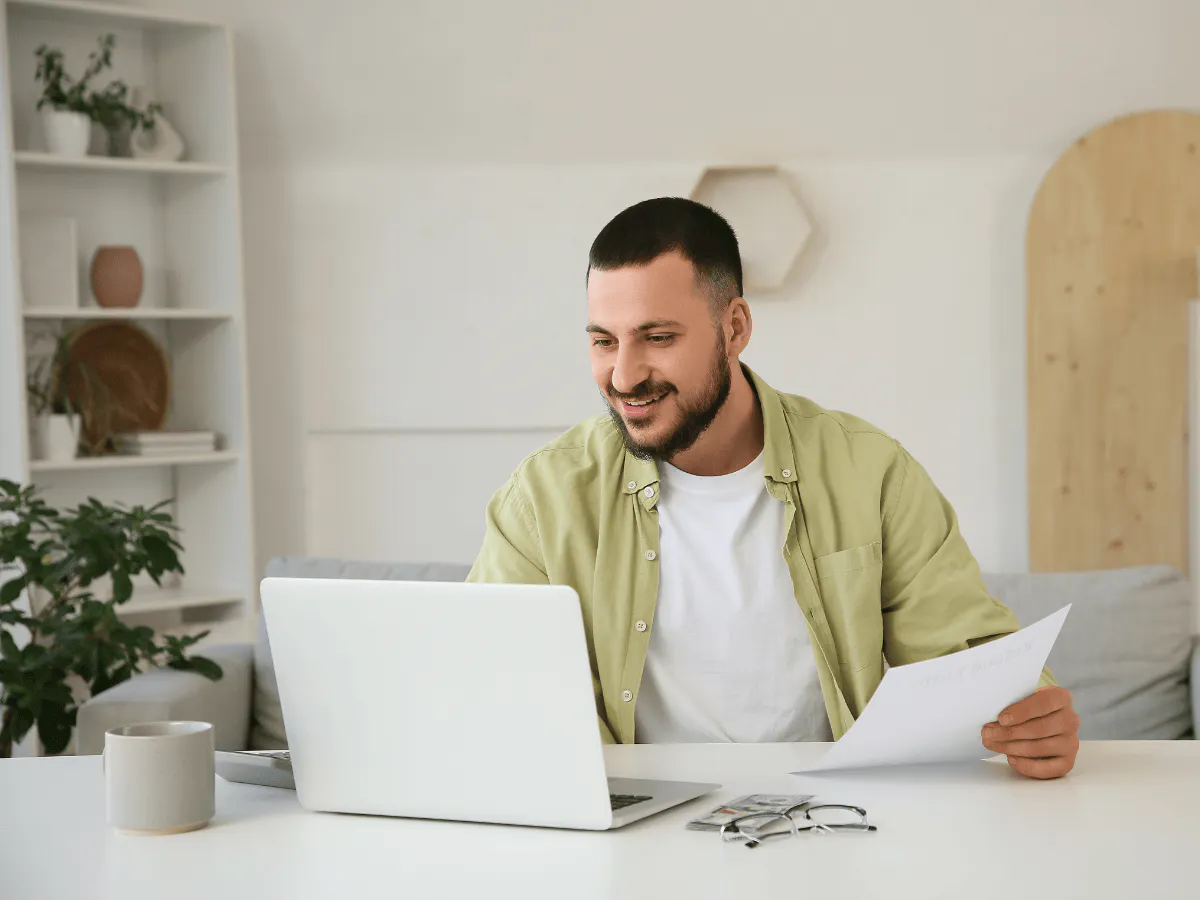 A person wearing a light green shirt sits at a desk with a laptop and papers, looking at the screen and smiling.
