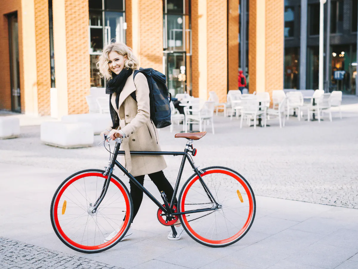 Een vrouw met blonde, warrige krullen en een stralende glimlach loopt met haar stijlvolle fixed-gear-fiets met rode velgen door een moderne kantoordistrict.