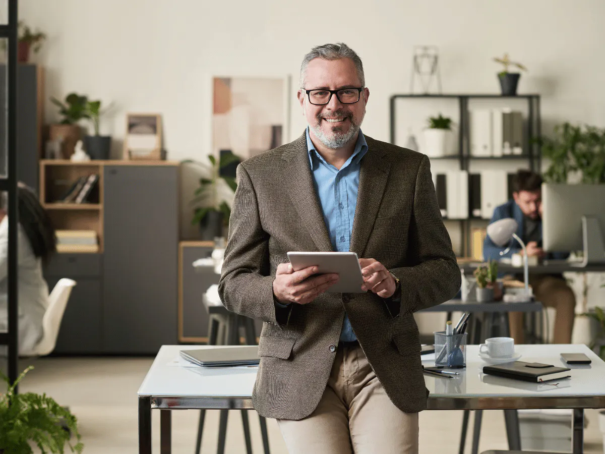 Een man in een bruine blazer staat in een licht kantoor met een tablet in zijn hand, leunt tegen een bureau en glimlacht naar de camera.