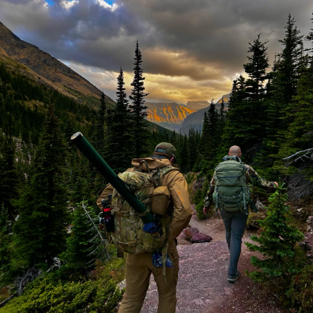 Two backpackers hike a trail through a dense forest in a mountainous landscape at dusk.