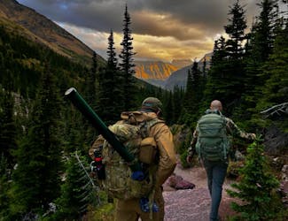 Two backpackers hike a trail through a dense forest in a mountainous landscape at dusk.