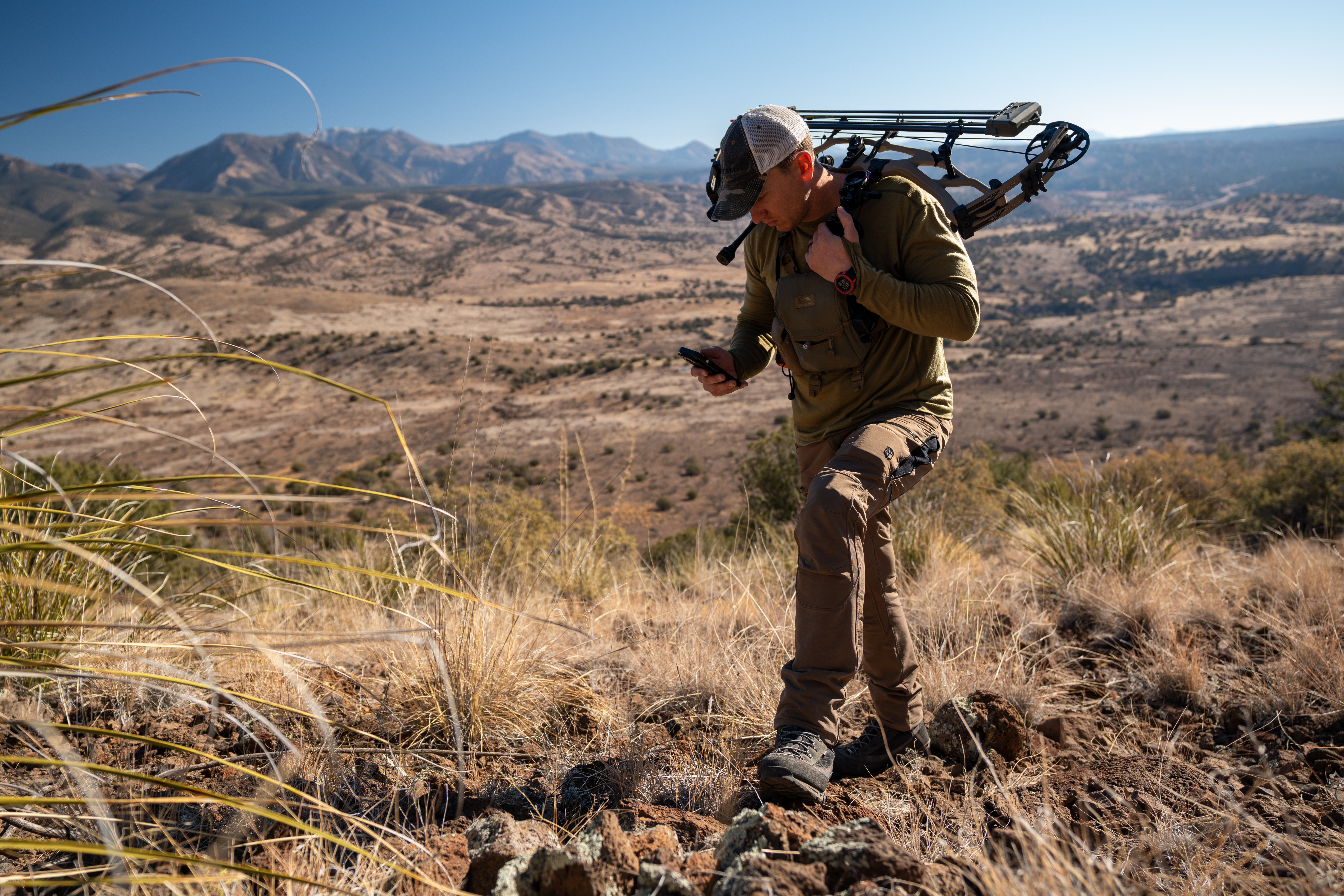Hunter with a bow and arrow looks at a handheld device in a dry, mountainous landscape.