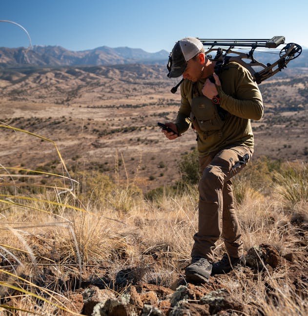 Hunter with a bow and arrow looks at a handheld device in a dry, mountainous landscape.