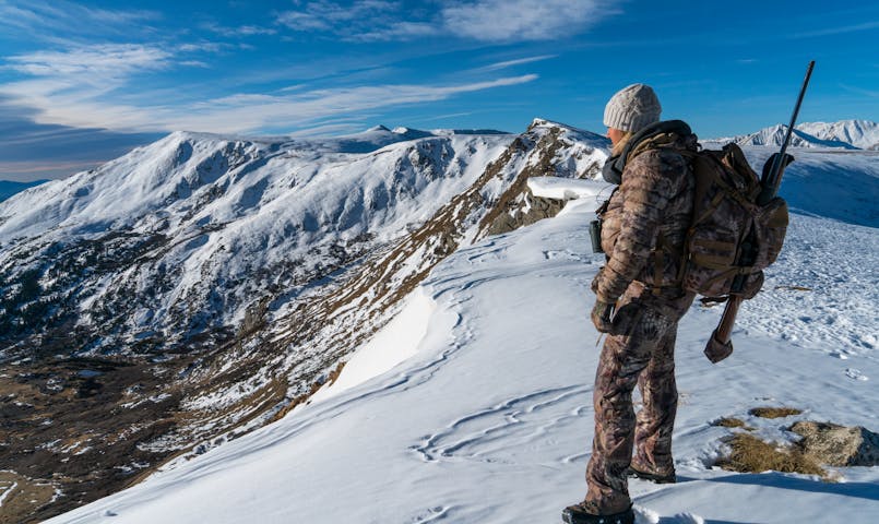 An individual dressed for cold-weather hunting, equipped with a rifle and backpack, observes a sprawling landscape of snow-covered peaks and valleys beneath a bright, partly cloudy sky.