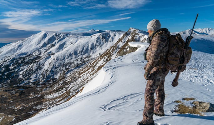 An individual dressed for cold-weather hunting, equipped with a rifle and backpack, observes a sprawling landscape of snow-covered peaks and valleys beneath a bright, partly cloudy sky.