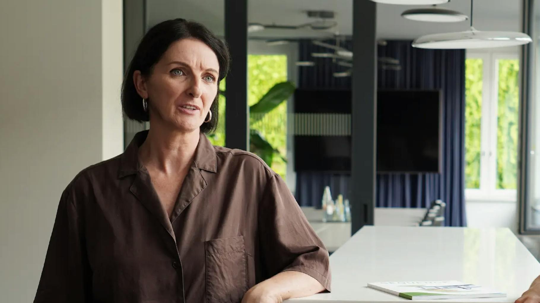 An employee of an advertising agency leans against a table and listens to someone.