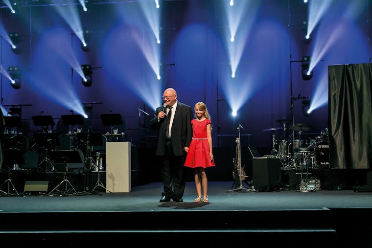 Gehard Schubert hät eine Rede auf der Bühne eines corporate events, neben ihm steht ein Mädchen in einem roten Kleid.