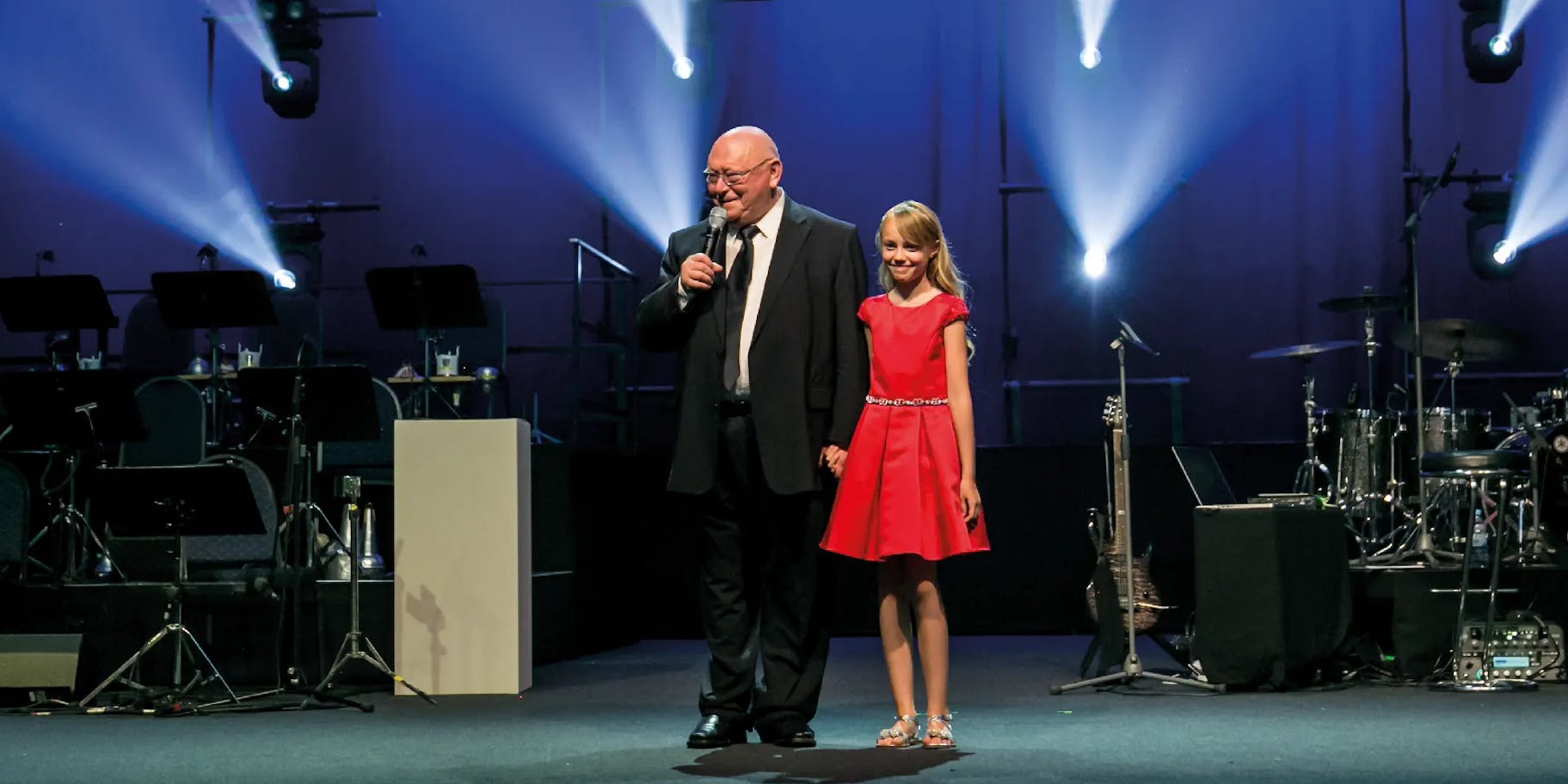 Gehard Schubert hät eine Rede auf der Bühne eines corporate events, neben ihm steht ein Mädchen in einem roten Kleid.
