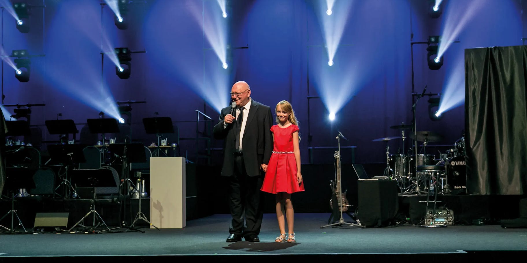 Gerhard Schubert is giving a speech on stage at a corporate event, with a girl in a red dress standing next to him.